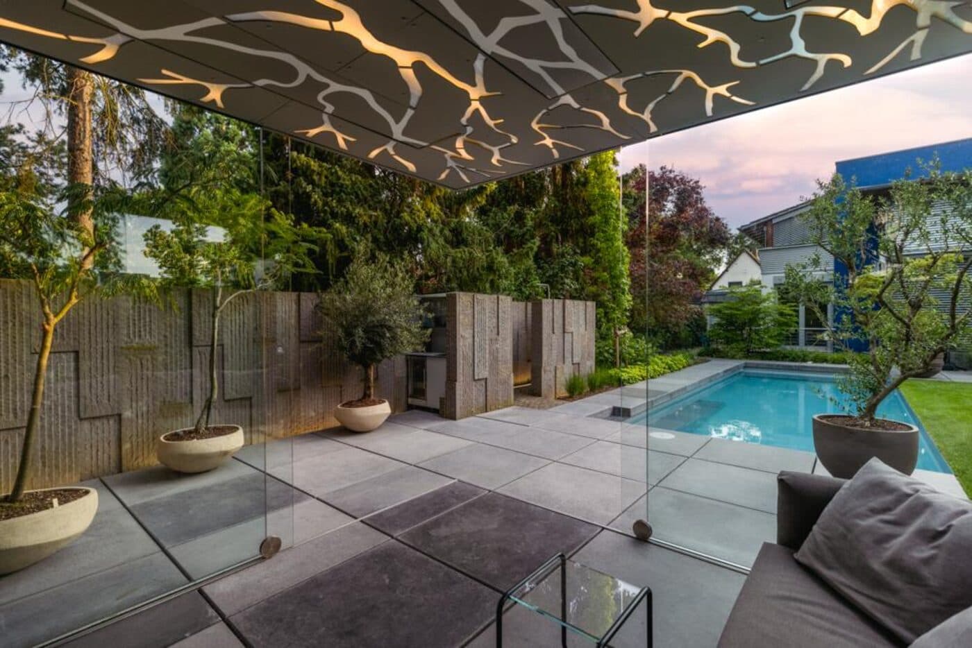 Moderne Gartenterrasse mit Glasüberdachung, beleuchteter Lichtinstallation, Pool und Natursteinplatten im Abendlicht