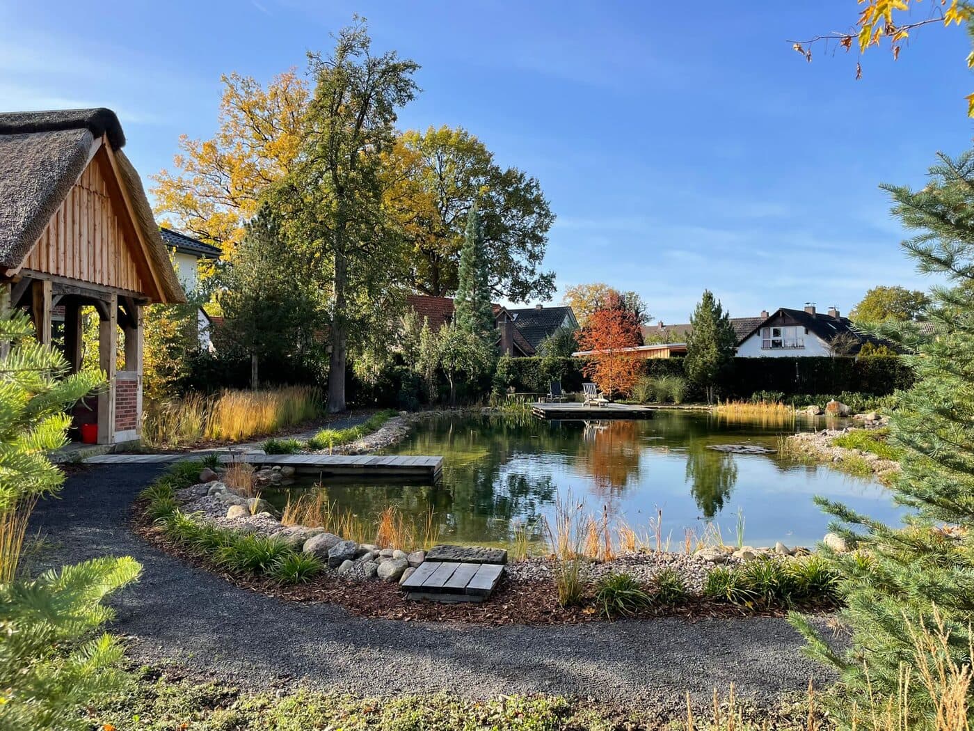 Naturnaher Schwimmteich mit reetgedecktem Gartenpavillon und herbstlicher Bepflanzung