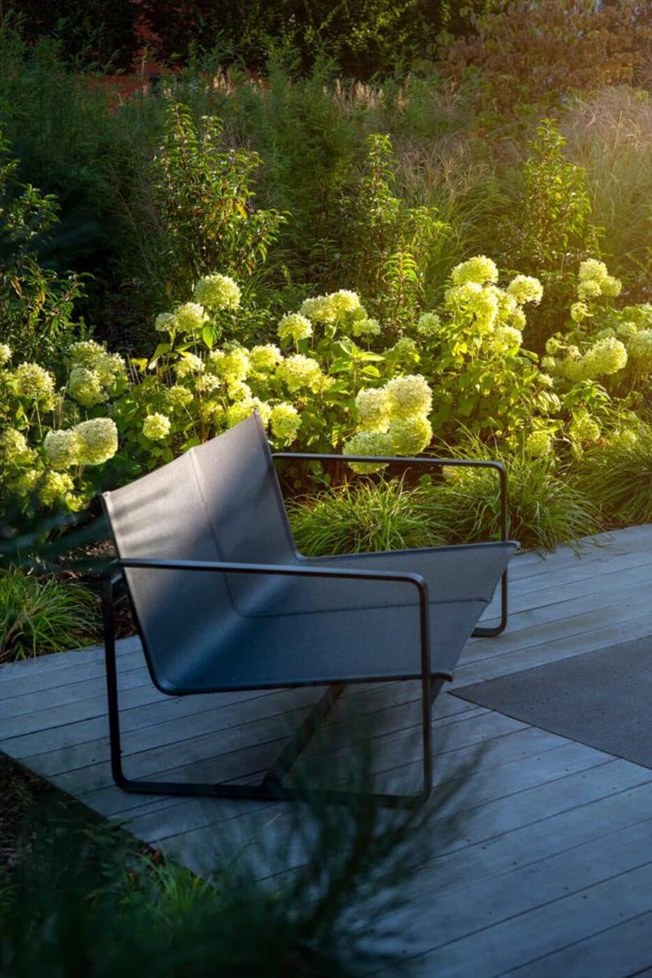 Loungebereich mit Designsessel auf Holzterrasse vor blühenden Hortensien im Abendlicht